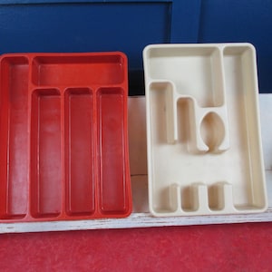 May include: Two plastic cutlery trays. One is red with rectangular compartments, the other is off-white with various shaped compartments. Both are designed to organise silverware and utensils. The trays are placed on a white shelf.