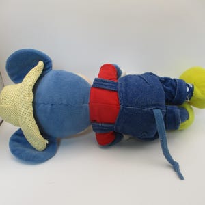May include: A plush toy mouse in a lying position. The mouse has a blue body, yellow ears, and a yellow hat. It wears red shorts and denim overalls. The mouse's tail is blue, and its feet are yellow.