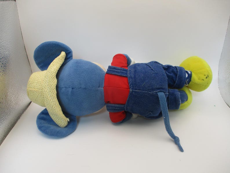 May include: A plush toy mouse in a lying position. The mouse has a blue body, yellow ears, and a yellow hat. It wears red shorts and denim overalls. The mouse's tail is blue, and its feet are yellow.
