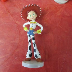May include: A figurine of the character Jessie from Toy Story. She wears a white shirt, blue jeans, and cow-print pants. Her red hat has a white trim. The figurine stands on a white base.