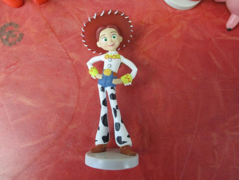 May include: A figurine of the character Jessie from Toy Story. She wears a white shirt, blue jeans, and cow-print pants. Her red hat has a white trim. The figurine stands on a white base.
