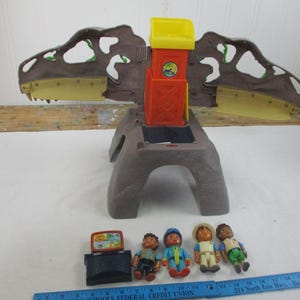 May include: A dinosaur-shaped playset with a brown and gray base, a yellow and orange tower, and a pair of opening jaws. Includes four small figurines and a black television. The playset is designed for children.
