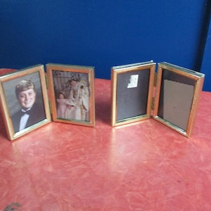 May include: Four gold-framed folding picture frames. Two frames contain photos, one of a person in a tuxedo and the other of a group of people. The other two frames are empty. The frames are on a red surface with a blue background.