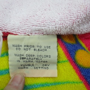 May include: Close-up of a colorful towel with a tag. The towel features pink, yellow, blue, and red sections. The tag provides washing instructions: wash prior to use, do not bleach, wash deep colors separately in warm water, tumble dry warm setting.