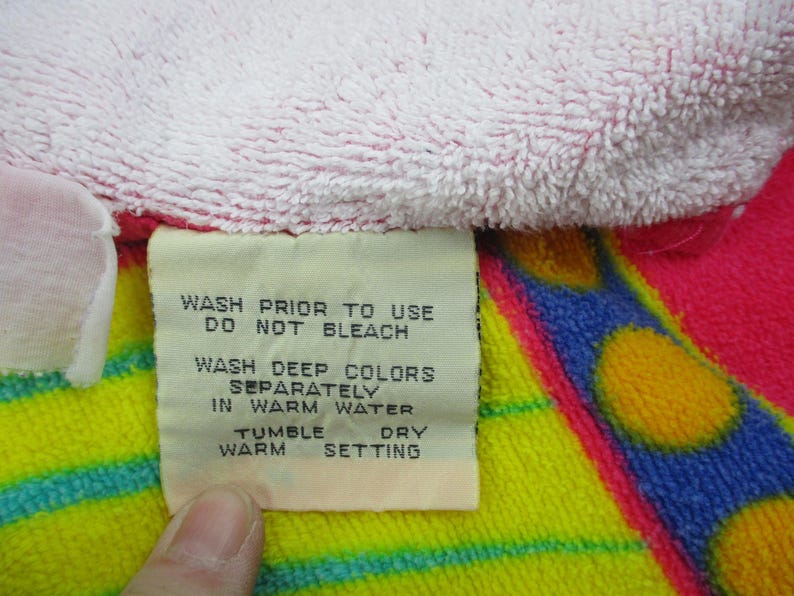 May include: Close-up of a colorful towel with a tag. The towel features pink, yellow, blue, and red sections. The tag provides washing instructions: wash prior to use, do not bleach, wash deep colors separately in warm water, tumble dry warm setting.