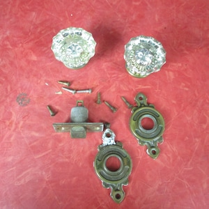 May include: Two clear, faceted glass doorknobs with brass hardware and screws, along with two brass escutcheons and a latch plate, arranged on a red surface. The hardware appears to be vintage.