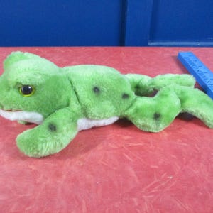 May include: A plush green frog toy with black spots and yellow eyes. The frog is lying on a red surface with a blue ruler in the background. The toy is made of soft, fuzzy material.