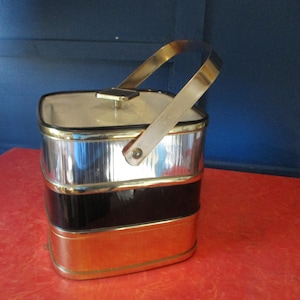 May include: Vintage metal ice bucket with a gold-tone handle and trim. The bucket has a rectangular shape with three sections: silver, black, and gold. The lid is cream-colored with a small gold knob. The handle is arched and attached to the top.