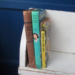 May include: A collection of vintage books, including "Cherry Ames, Clinic Nurse" and "Cherry Ames, Student Nurse." The books are in various colors, with the covers showing signs of wear. The books are standing upright on a white surface.