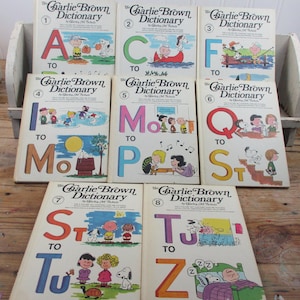 May include: A collection of eight vintage "The Charlie Brown Dictionary" books. Each book features a large letter of the alphabet, with colorful illustrations of Charlie Brown, Snoopy, and other Peanuts characters. The books are arranged on a wooden surface.