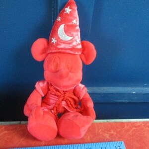 May include: A red plush Mickey Mouse figure dressed as a sorcerer, wearing a pointed hat with a crescent moon and star accents. The figure has large ears and is seated, with a red outfit and a rope-like tie.