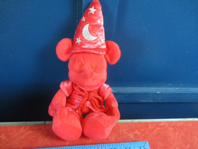 May include: A red plush Mickey Mouse figure dressed as a sorcerer, wearing a pointed hat with a crescent moon and star accents. The figure has large ears and is seated, with a red outfit and a rope-like tie.