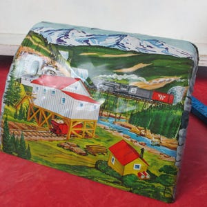 May include: A vintage tin box with a colorful landscape scene. The artwork depicts a train on a bridge, a building with a red roof, and a river flowing through a valley. Mountains are in the background.