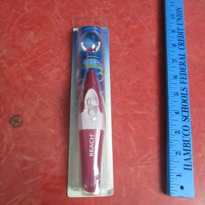 May include: A new Reach Access toothbrush in its original packaging. The toothbrush is a combination of white and burgundy colors. A blue ruler is next to the toothbrush.