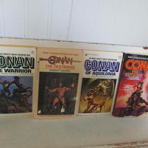 Könnte beinhalten: Vier Vintage-Taschenbücher aus der Conan der Barbar-Reihe. Die Cover zeigen Actionszenen mit Titeln wie "Conan der Krieger" und "Conan der Zerstörer". Die Bücher sind auf einer weißen Oberfläche angeordnet.