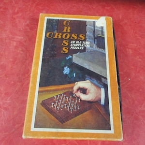 May include: Vintage "Cross" puzzle game box with an orange border. The box features an image of the game in play, with a hand placing a peg. The text on the box reads "Cross, An Old Time Stimulating Puzzler."