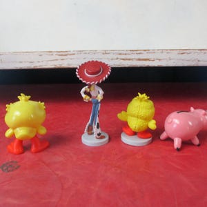 May include: Four colorful character figurines on a red surface. The figurines include a yellow chick with orange feet, a pink pig, a cowboy figure with a red hat, and another yellow chick with orange feet. The figurines are likely from a children's movie.