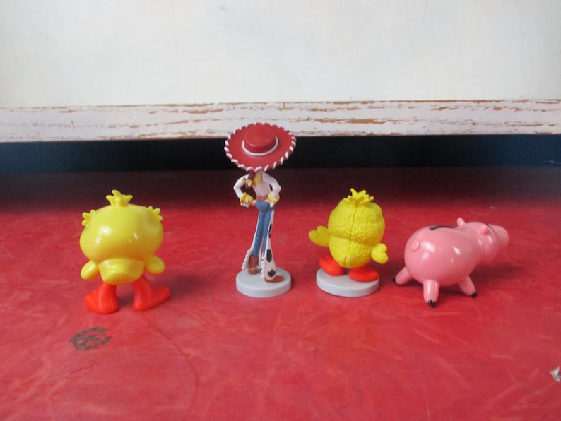 May include: Four colorful character figurines on a red surface. The figurines include a yellow chick with orange feet, a pink pig, a cowboy figure with a red hat, and another yellow chick with orange feet. The figurines are likely from a children's movie.