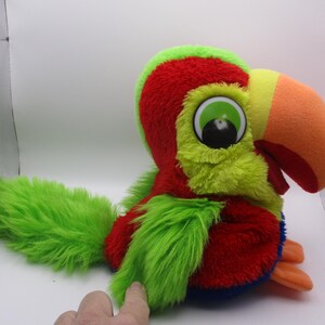 May include: A plush parrot toy featuring bright red, lime green, and blue fur. The bird has a large, soft orange beak, a lime green crest, and oversized white eyes with black pupils. It is depicted in a seated position with orange feet.