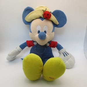 May include: A plush Mickey Mouse toy wearing a straw hat with a red ladybug and blue overalls. The toy has white gloves, yellow feet, and blue ears. The toy is sitting on a white surface.