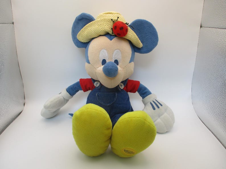 May include: A plush Mickey Mouse toy wearing a straw hat with a red ladybug and blue overalls. The toy has white gloves, yellow feet, and blue ears. The toy is sitting on a white surface.