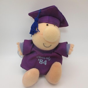 May include: A soft toy doll dressed in a purple graduation outfit, including a cap and gown. The doll has a large nose and a smiling expression. The gown is embroidered with "Class of '84". The cap has a blue tassel.