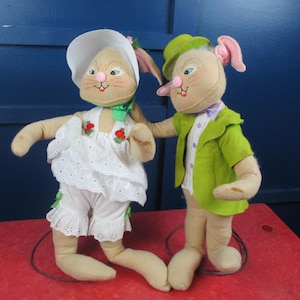 May include: Two plush bunny dolls. One wears a white dress with red rose accents and a matching hat. The other doll wears a green jacket and hat with a purple bow tie. Both dolls have long ears and are standing on a red surface.