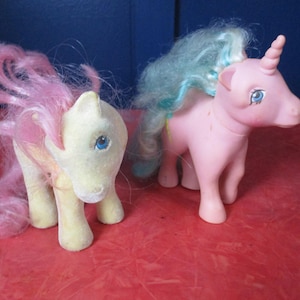 Könnte beinhalten: Zwei Vintage-Spielzeugponys. Eines ist hellgelb mit rosa Haaren, das andere ist rosa mit blau-gelben Haaren und einem Horn. Beide haben blaue Augen und stehen auf einer roten Oberfläche. Die Ponys sind ein nostalgisches Spielzeug.