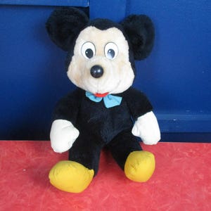 Op de afbeelding: Een pluche Mickey Mouse pop, met een zwart lichaam, grote zwarte oren en een cr&egrave;mekleurig gezicht. De pop heeft een blauwe vlinderdas, witte handschoenen en gele voeten. De achtergrond is een blauwe muur en een rood oppervlak.