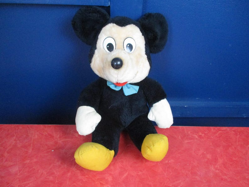 Op de afbeelding: Een pluche Mickey Mouse pop, met een zwart lichaam, grote zwarte oren en een cr&egrave;mekleurig gezicht. De pop heeft een blauwe vlinderdas, witte handschoenen en gele voeten. De achtergrond is een blauwe muur en een rood oppervlak.