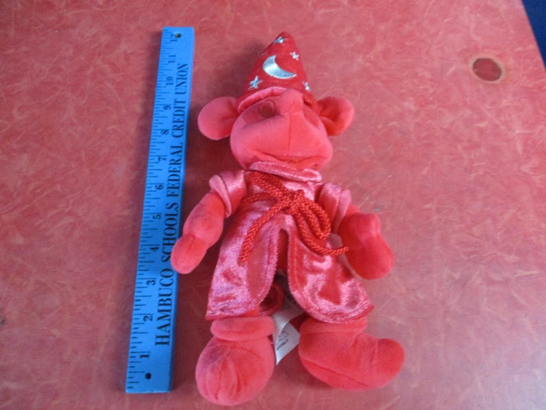 May include: A plush red wizard doll with a pointed hat adorned with a crescent moon and stars. The doll is wearing a red robe and boots. A blue ruler, approximately 8 inches long, is next to the doll.