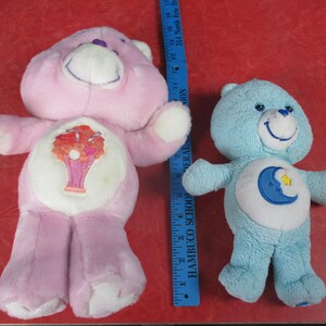 May include: Two plush Care Bears. One is light pink with a tree design on its belly, the other is light blue with a moon and star design. A blue ruler is next to the bears.