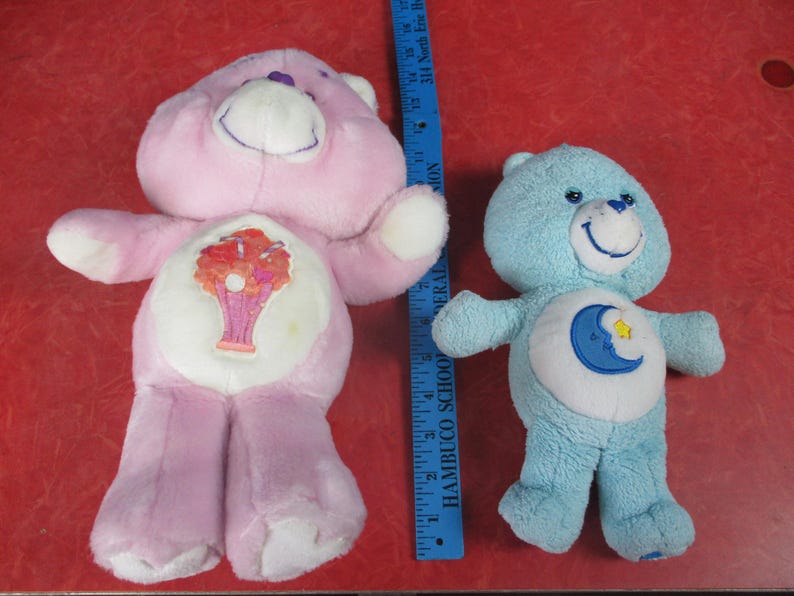 May include: Two plush Care Bears. One is light pink with a tree design on its belly, the other is light blue with a moon and star design. A blue ruler is next to the bears.