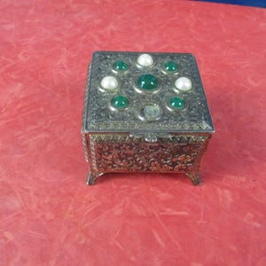 May include: A small, ornate silver-colored box with a hinged lid. The lid is decorated with green and white gemstones. The box has a detailed floral pattern on the sides and rests on small feet. A decorative jewelry box.