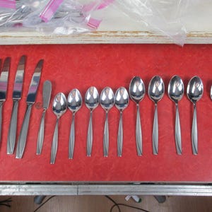 May include: A set of stainless steel cutlery, including knives, spoons, and a butter knife, displayed on a red surface. The cutlery has a sleek, modern design. The spoons are of varying sizes.