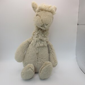 May include: A plush llama toy in a seated position. The llama is a light beige color with a fluffy mane and neck. It has small, rounded ears and a gentle expression. The toy is soft and cuddly, perfect for children.