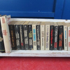 May include: A collection of vintage books by Lawrence Sanders, displayed on a white shelf against a blue background. The book spines are in various colors, with titles like "The Anderson Tapes" and "Capital Crimes".