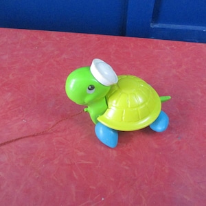 May include: A colorful toy turtle with a green head, yellow shell, and blue feet. The turtle wears a white hat and has a pull string attached. The toy is on a red surface.