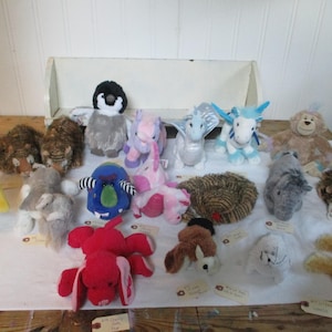 May include: A collection of plush toys, including a penguin, dragon, and lion, are arranged on a white surface. The toys are various colors, such as blue, pink, and brown. Some toys have tags attached.