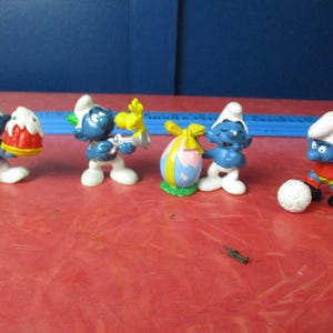 Puede incluir: Una colección de figuras de los Pitufos. Las figuras son azules con gorros blancos y pantalones blancos. Un Pitufo sostiene un pastel, otro un pájaro amarillo y otro un huevo de Pascua. Un cuarto Pitufo juega al fútbol. Sobre una superficie roja.