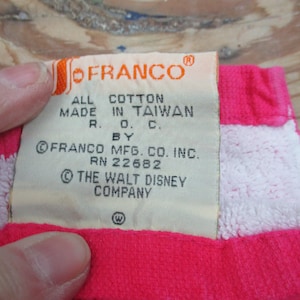 May include: A close-up of a tag from a vintage item. The tag is cream-colored with the "OFRANCO" logo in orange. Text on the tag reads "ALL COTTON MADE IN TAIWAN R.O.C. BY FRANCO MFG. CO. INC. RN 22682 &copy; THE WALT DISNEY COMPANY". The item is pink and white.