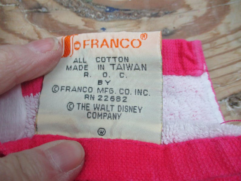 May include: A close-up of a tag from a vintage item. The tag is cream-colored with the "OFRANCO" logo in orange. Text on the tag reads "ALL COTTON MADE IN TAIWAN R.O.C. BY FRANCO MFG. CO. INC. RN 22682 &copy; THE WALT DISNEY COMPANY". The item is pink and white.