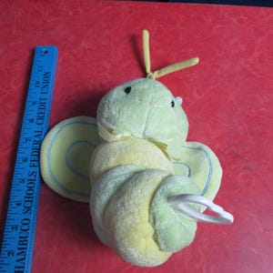 May include: A plush, light green and yellow butterfly-shaped toy with black eyes and yellow antennae. The toy has a white plastic ring attached. A blue ruler with white text is next to the toy.