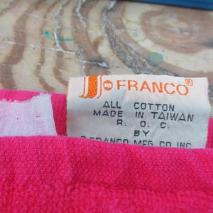 May include: Close-up of a bright pink textile with a white tag. The tag reads "JO FRANCO" and "ALL COTTON MADE IN TAIWAN". The textile is likely a towel or similar item.