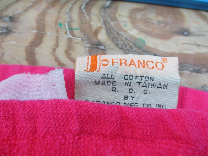 May include: Close-up of a bright pink textile with a white tag. The tag reads "JO FRANCO" and "ALL COTTON MADE IN TAIWAN". The textile is likely a towel or similar item.