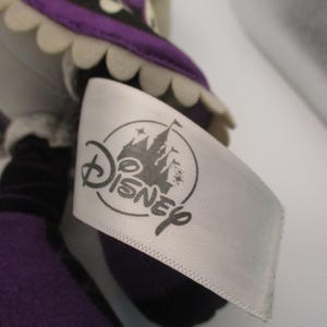 Puede incluir: Una mu&ntilde;eca de peluche con una etiqueta blanca con el logotipo de Disney. La mu&ntilde;eca tiene un atuendo morado y negro con un ribete blanco festoneado. La etiqueta es blanca con el logotipo de Disney en gris.