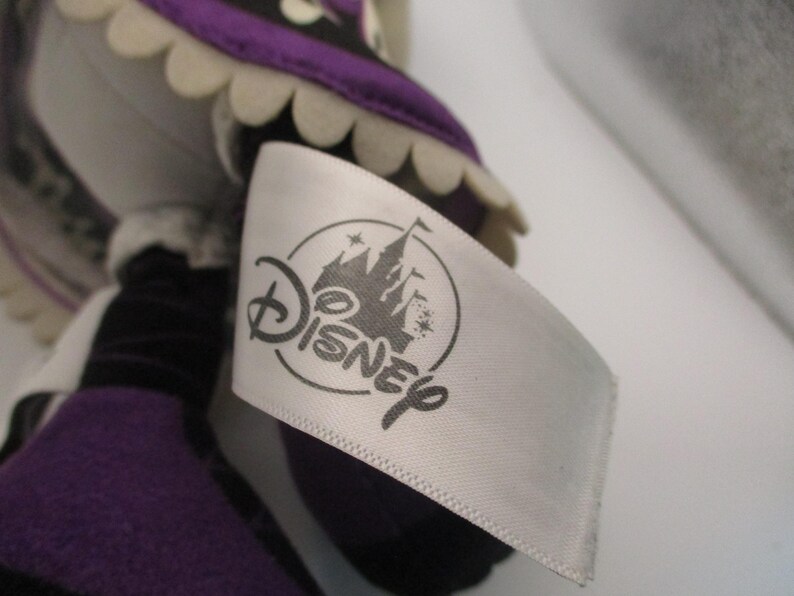 Puede incluir: Una mu&ntilde;eca de peluche con una etiqueta blanca con el logotipo de Disney. La mu&ntilde;eca tiene un atuendo morado y negro con un ribete blanco festoneado. La etiqueta es blanca con el logotipo de Disney en gris.