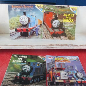 Puede incluir: Cuatro libros infantiles con Thomas el tanque. Los títulos incluyen "Edward's Exploit", "James in a Mess", "Trouble for Thomas" y "Danger at the Dieselworks". Los libros se muestran en una estantería blanca y una superficie roja.