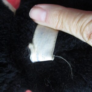 Op de afbeelding: Close-up van een zwart pluche speelgoed met een wit label. Het label is met een witte draad aan het pluche speelgoed bevestigd. Het pluche speelgoed is zacht en pluizig. Het label heeft tekst.