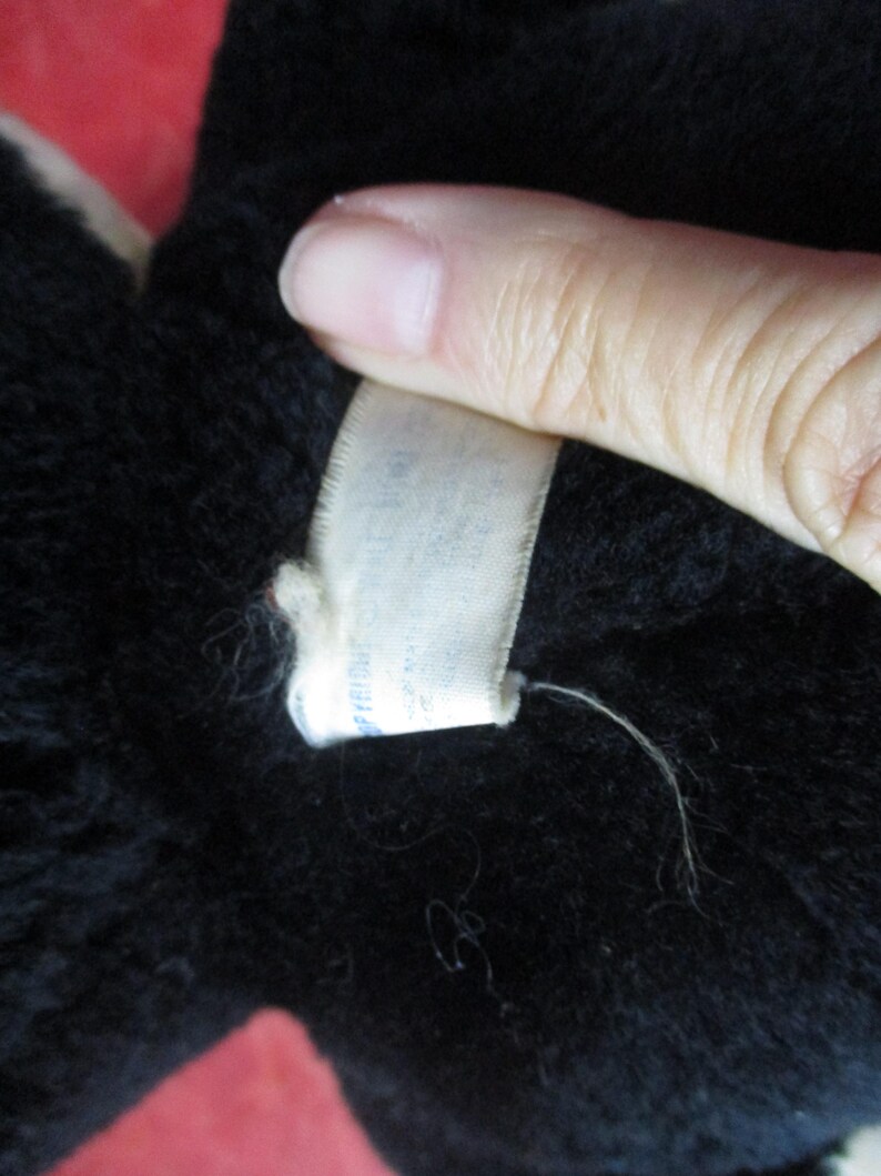 Op de afbeelding: Close-up van een zwart pluche speelgoed met een wit label. Het label is met een witte draad aan het pluche speelgoed bevestigd. Het pluche speelgoed is zacht en pluizig. Het label heeft tekst.